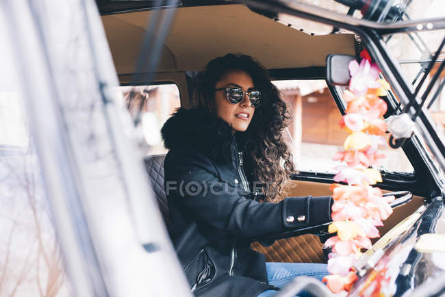 Mujer joven con abrigo negro y gafas de sol sentado dentro del coche - foto de stock