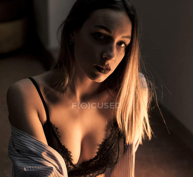 Encantadora joven en sujetador de encaje negro y camisa mirando a la cámara mientras está sentada en la habitación oscura - foto de stock