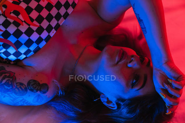 Молода жінка з рукою на лобі, дивлячись геть, лежачи в кімнаті з червоним і синім світлом — стокове фото
