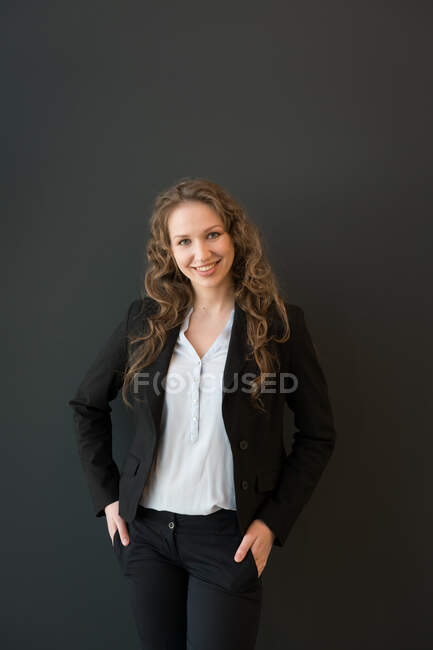 Beautiful blond young woman in dark elegant suit standing confidently with crossed arms and looking at camera smiling on black background — Stock Photo