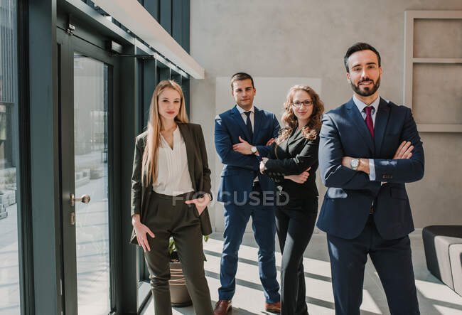 Group of office colleagues in elegant dark suits standing together relaxed with crossed arms and looking at camera smiling in spacious room at glass wall and door — Stock Photo