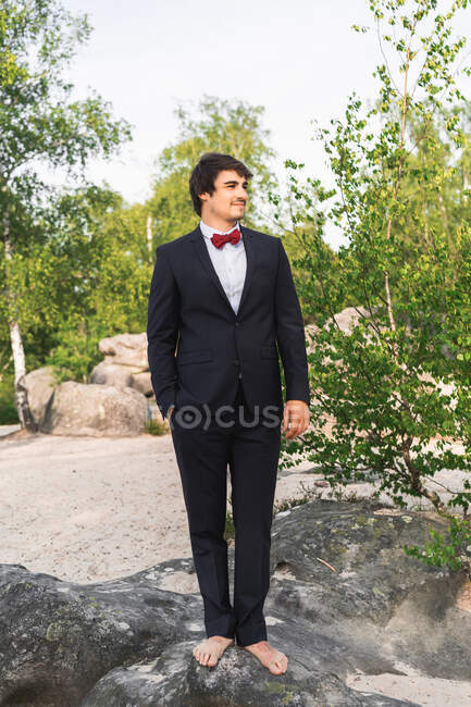 Jovem sorrindo descalço vestindo terno preto com gravata borboleta vermelha e em pé na rocha do litoral olhando sonhadoramente longe — Fotografia de Stock