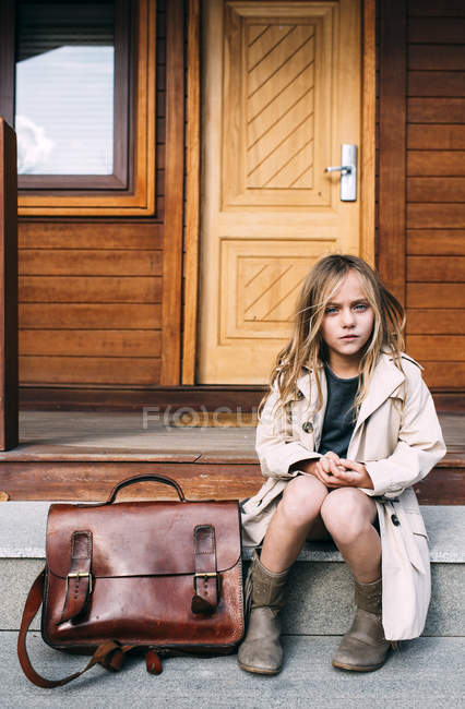 Blond girl in trench-coat with leather briefcase sitting in front of wooden house and looking at camera — Stock Photo
