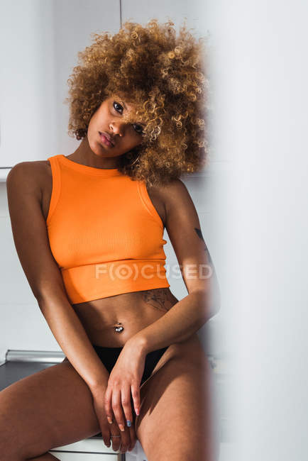 Sensual joven mujer en naranja superior sentado en el mostrador y mirando a la cámara - foto de stock