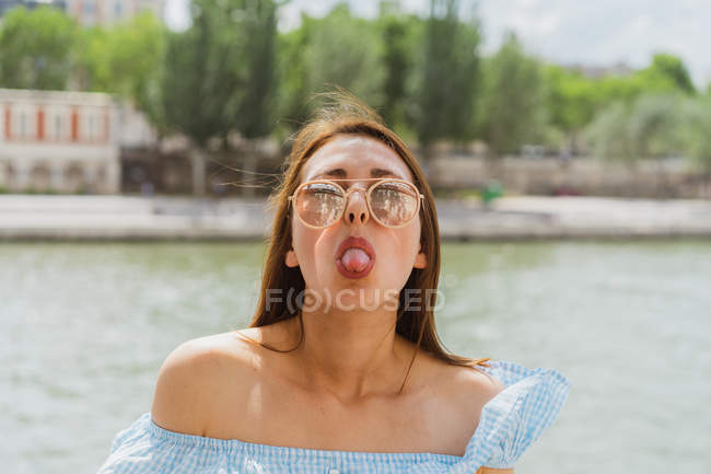 Jolie femme en lunettes de soleil montrant la langue sur le front de mer sur fond flou — Photo de stock