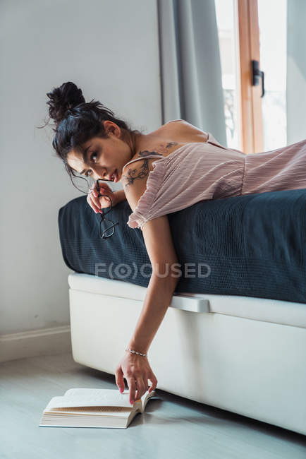 Кокетливая женщина лежит на кровати с книгой на полу и кусает очки, соблазнительно глядя в камеру — стоковое фото