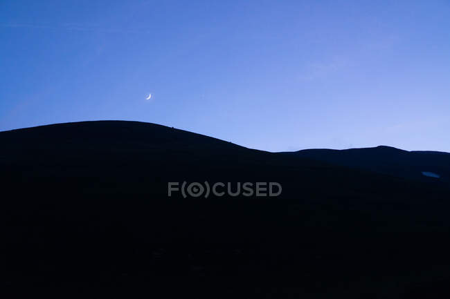 Мінімалістичний пейзаж чорного силуету гірських пагорбів на тлі сутінкового блакитного неба з півмісяцем — стокове фото