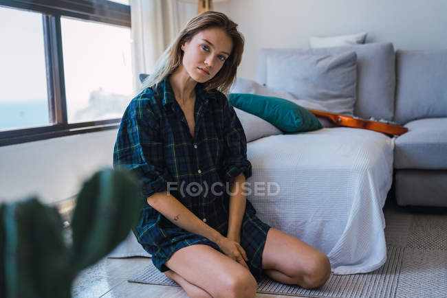 Mujer joven con camisa de gran tamaño a cuadros sentada en el suelo cerca del sofá - foto de stock