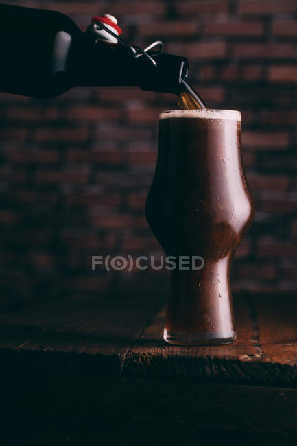Pouring stout beer in glass from bottle on dark wooden table — Stock Photo
