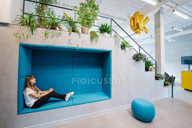 Вид збоку жінки відпочиває в синій затишній ніші в офісному коридорі, використовуючи смартфон у дозвіллі — стокове фото