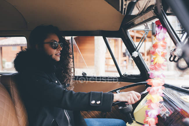 Mujer joven con abrigo negro y gafas de sol conduciendo coche retro - foto de stock