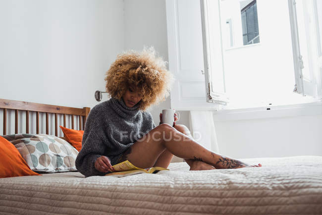 Seducente donna etnica in maglione seduta sul letto con tazza di caffè e rivista di lettura alla luce del giorno — Foto stock