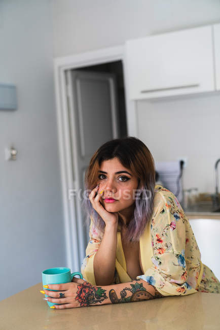 Mujer joven en bata de seda sosteniendo taza con bebida caliente y mirando a la cámara mientras se apoya en la mesa en la cocina - foto de stock