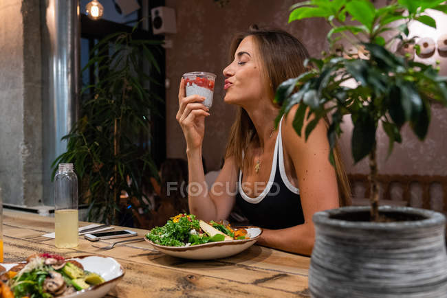 Mujer joven sentada a la mesa de madera con comida y sosteniendo una taza de postre de plástico - foto de stock