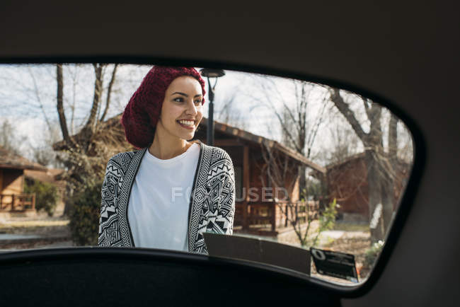 Jovem mulher de chapéu de lã em pé atrás do carro no campo — Fotografia de Stock