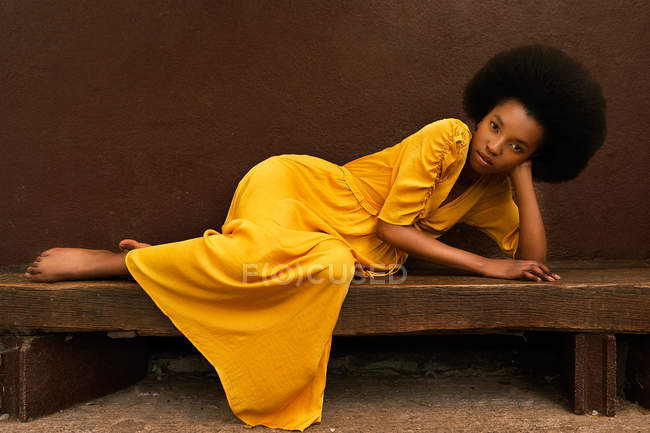 Retrato de mulher afro-americana em amarelo brilhante vestido longo deitado no banco de madeira no fundo marrom — Fotografia de Stock