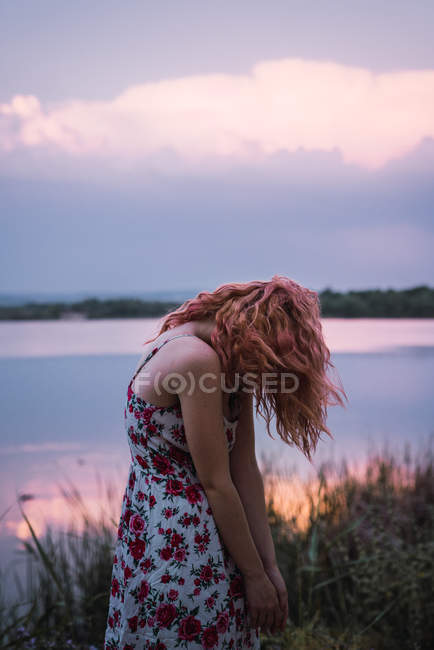Frau im Kleid mit haarsträubendem Gesicht steht bei Sonnenuntergang am Seeufer — Stockfoto