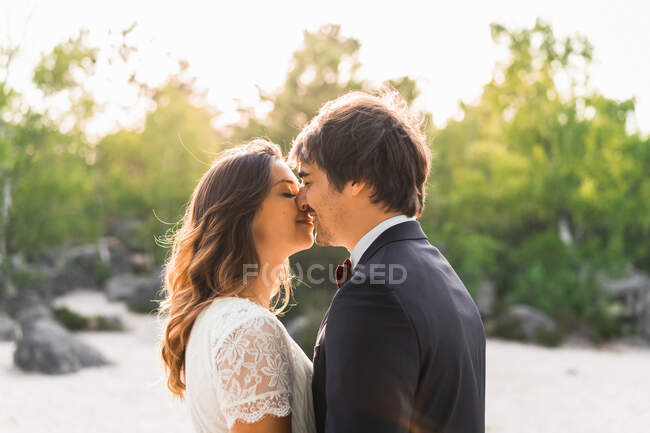 Casal em vestidos de casamento em pé na rocha e abraçando alegremente contra árvores verdes e céu azul — Fotografia de Stock