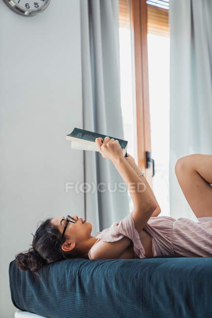 Junge brünette Frau liest auf dem Bett, während sie zu Hause auf dem Bett liegt — Stockfoto