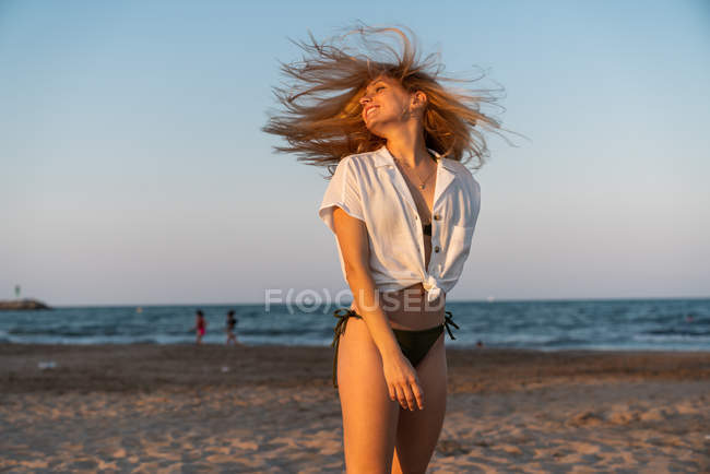 Jeune femme en bikini et chemise avec des cheveux soufflants debout sur la plage au coucher du soleil — Photo de stock