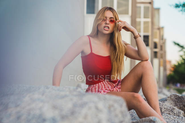 Young woman in red shorts and tank top and grimacing while sitting on rocks on street — Stock Photo