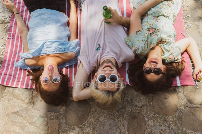 Mulheres jovens e homem em óculos de sol e roupas casuais sorrir enquanto deitado em cobertor ao ar livre — Fotografia de Stock
