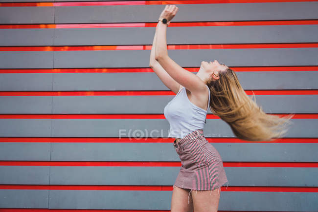 Jovem mulher em roupa casual rindo e sacudindo o cabelo enquanto está de pé contra a parede listrada — Fotografia de Stock