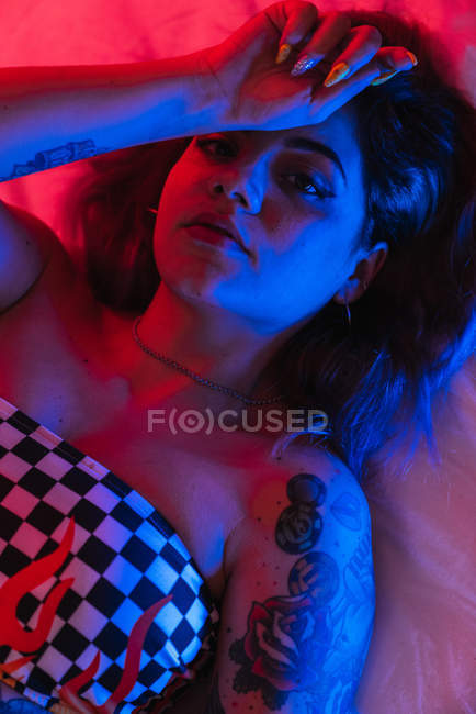 Молодая женщина с рукой на лбу смотрит в камеру, лежа в комнате с красным и синим светом — стоковое фото