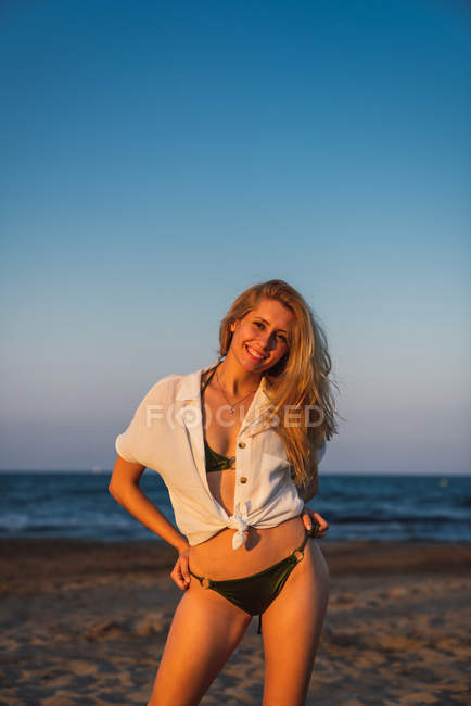 Relaxed smiling woman in bikini and shirt standing on beach at sunset — Stock Photo
