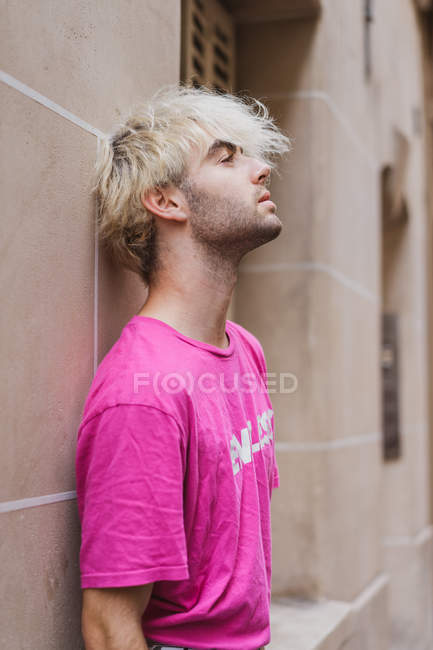Stylish blond man in pink t-shirt leaning on building wall on street — Stock Photo