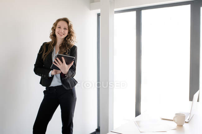 Elegant woman wearing suit and using tablet while standing in bright daylight inside of modern office — Stock Photo