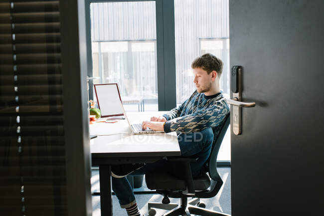 Вид сбоку мужчины в повседневной одежде, удобно сидящего на офисном стуле и печатающего на клавиатуре ноутбука на белом столе с большим окном на заднем плане, сделанным через открытую дверь — стоковое фото