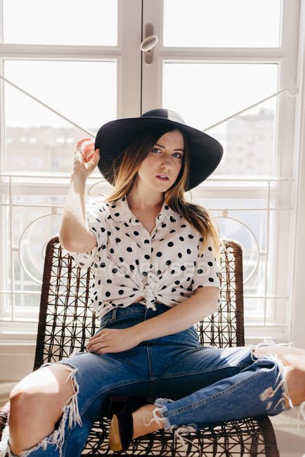 Stylische junge Frau mit Hut sitzt auf Stuhl und blickt in die Kamera — Stockfoto