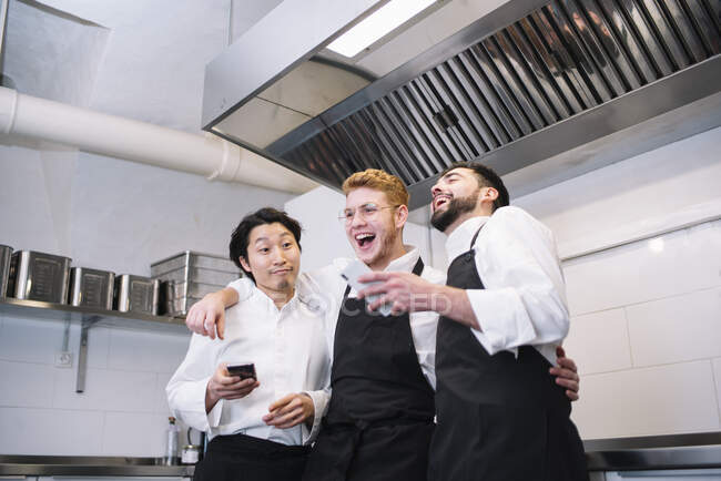 Trois hommes divers en uniforme de cuisine riant et naviguant smartphones tout en se tenant sur la cuisine du restaurant ensemble — Photo de stock