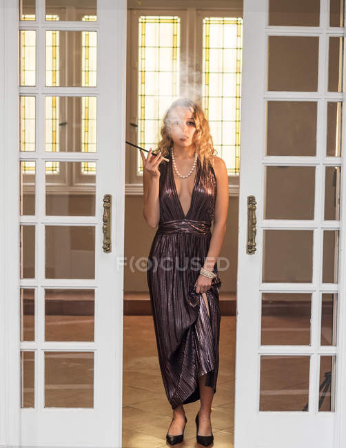 Élégante jeune femme en robe de soirée fumer cigarette tout en se tenant à la porte de la maison de luxe — Photo de stock