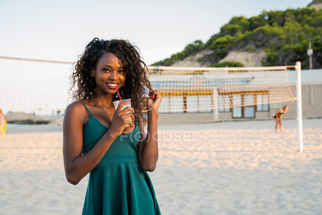 Elegante mujer negra con bebida de pie en la playa y mirando a la cámara - foto de stock