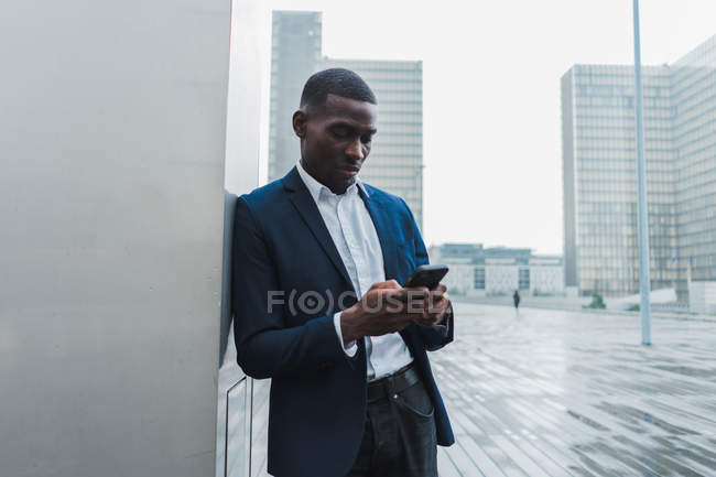 Hombre joven en traje elegante de pie apoyado en la pared del edificio y el uso de teléfono móvil - foto de stock