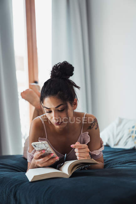 Pretty brunette woman relaxing lying on bed with book opened and using smartphone — Stock Photo