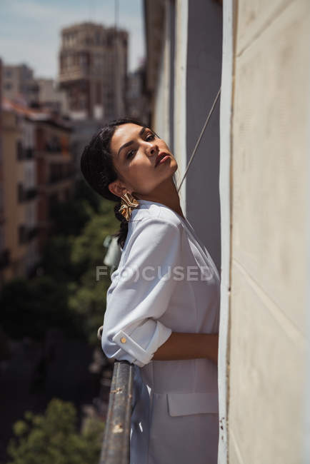 Sensual mulher morena em elegante jaqueta branca e brincos dourados olhando para a câmera enquanto se inclina na cerca da varanda — Fotografia de Stock