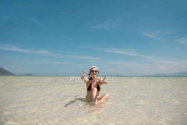 Cheerful woman gesturing thumbs up and sitting in ocean water — Stock Photo