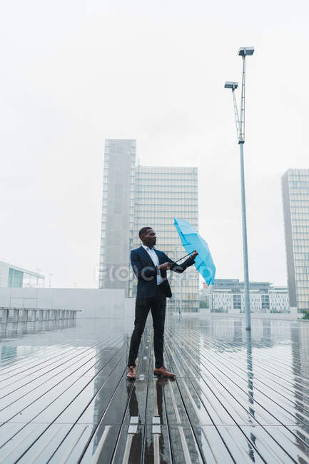 Uomo d'affari etnico con l'ombrello blu in piedi sul marciapiede bagnato della città — Foto stock