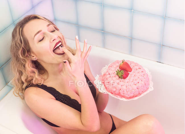 Sinnliche Dame in schwarzer Unterwäsche sitzt in der Badewanne und schmeckt süße Sahne aus leckerem Kuchen — Stockfoto