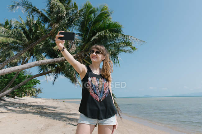 Cheerful woman in sunglasses taking selfie at palm tree on beach — Stock Photo