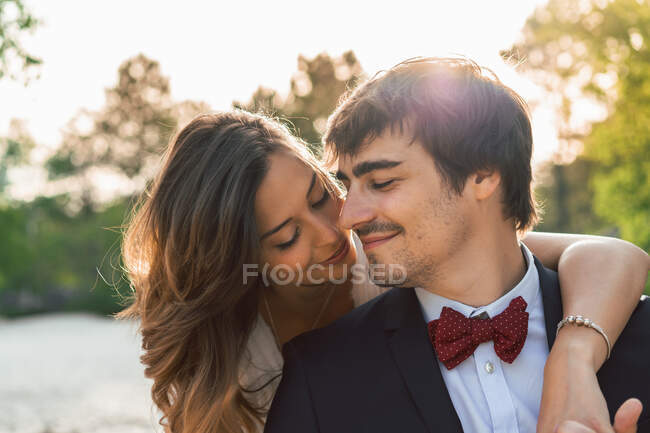 Feliz elegante hombre y mujer en trajes de novia abrazándose en la roca de la playa y mirando sonriente lejos a la luz del sol - foto de stock