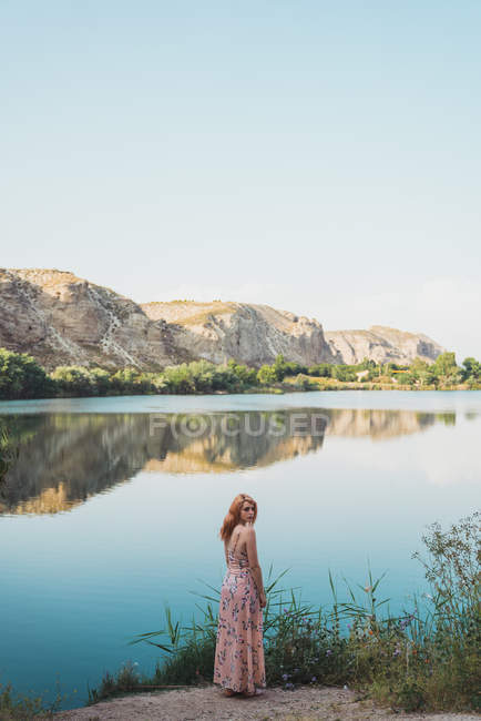 Женщина в длинном летнем платье стоит на берегу озера и смотрит через плечо — стоковое фото