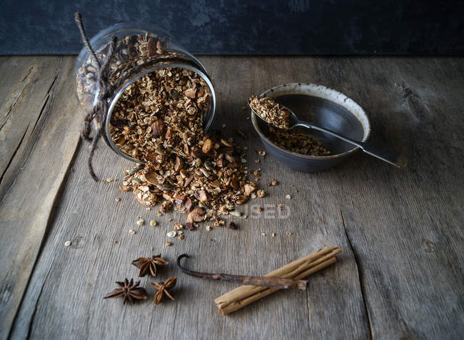 Pot renversé de délicieux granola chai-épicé sur table en bois avec bol et divers arômes — Photo de stock