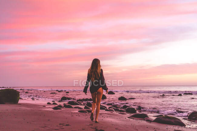 Vista posteriore della vestibilità sexy giovane femmina in camicia nera che cammina sulla riva dell'oceano con sabbia e rocce al tramonto con bel cielo nuvoloso rosa sullo sfondo — Foto stock