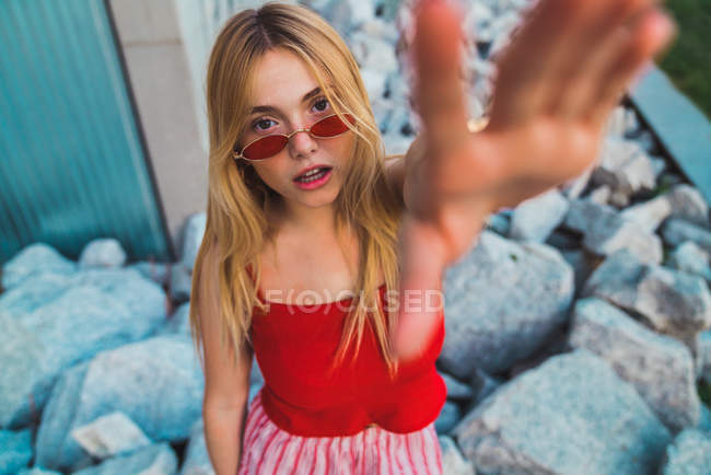 Красивая молодая женщина в стильных солнцезащитных очках протягивает руку к камере, стоя на скалах на открытом воздухе — стоковое фото