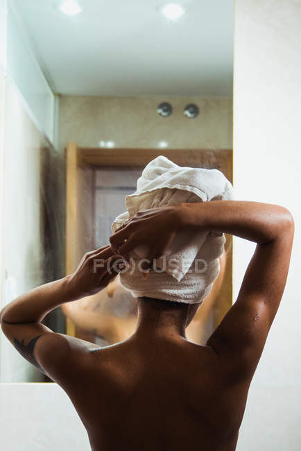 Topless ethnic woman wrapping towel on head after shower standing in front of mirror — Stock Photo