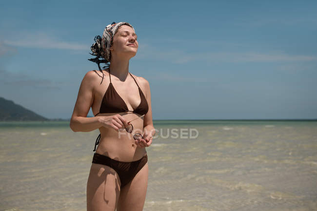 Woman in swimsuit and bandana standing on sandy coast — Stock Photo